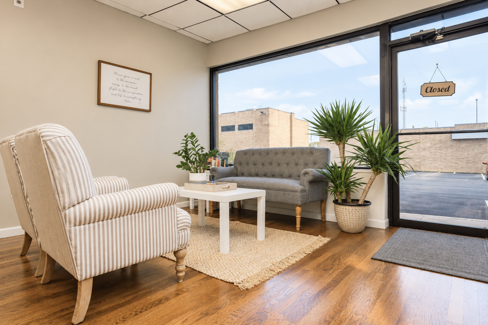 A cozy waiting area with beige and gray furniture, three potted plants, a small white table, a framed inspirational quote on the wall, and a large window with a door that has a closed 'Closed' sign, overlooking a parking lot and brick building.