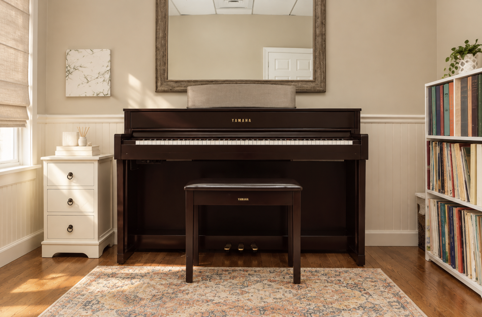 A room with a Yamaha upright piano, a matching bench, a small white side table on the left, and a bookshelf with colorful books on the right. There is a large mirror above the piano, a piece of wall art, and a window with beige shades on the left. The floor is wooden with a patterned rug.