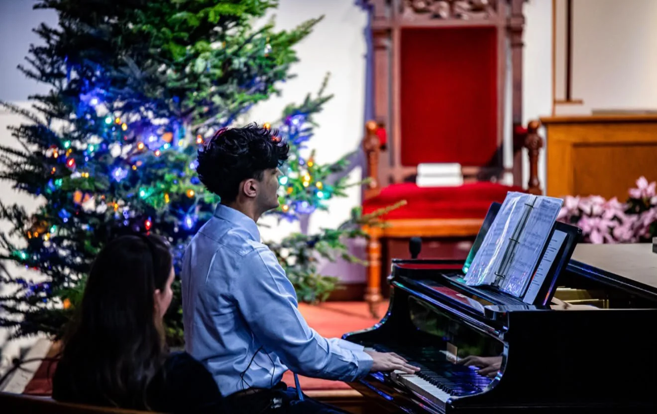 Two people sitting at a grand piano, with one playing and sheet music open on the music stand, beside a decorated Christmas tree with colorful lights, in a room with wooden furniture and pink flowers.
