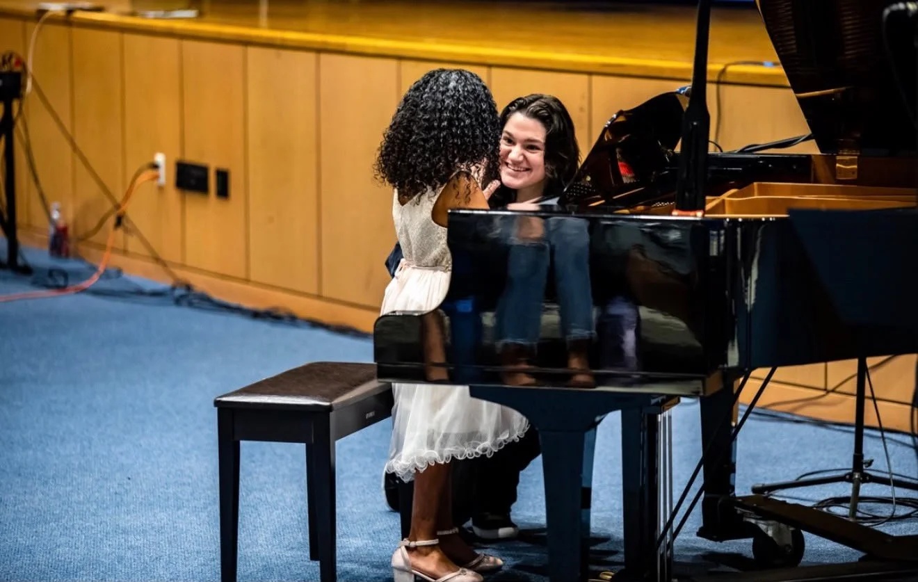 A young girl and a woman smiling at each other by a black grand piano in a room with wooden paneling and blue carpeting.