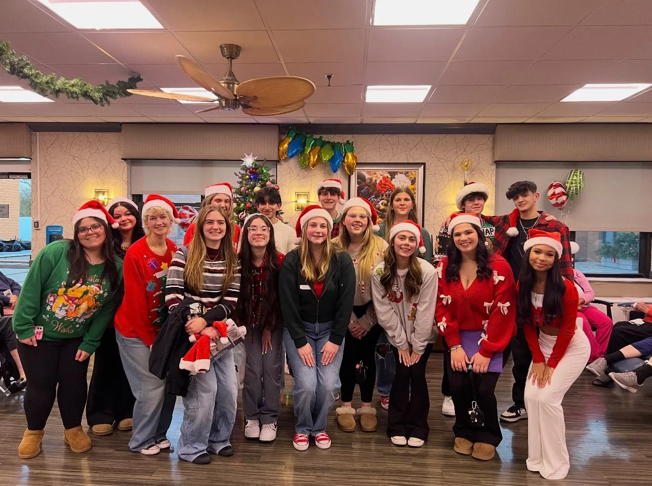 Group of teenagers dressed in Christmas sweaters and Santa hats Christmas decorations, tree, balloons, smiling at the camera.