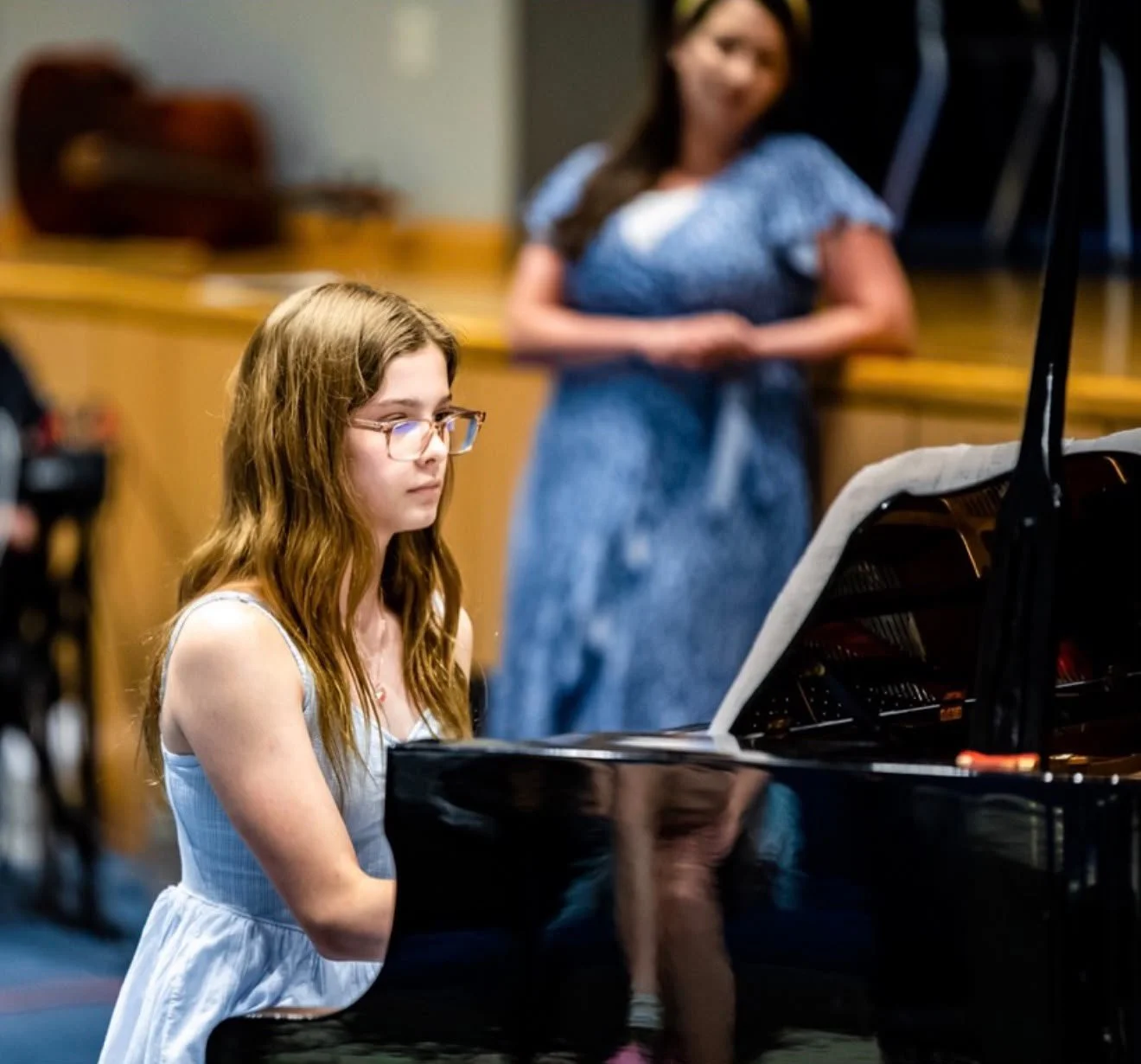 Young girl with brown hair and glasses playing a grand piano during a performance or practice session.