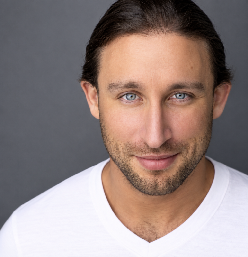 Close-up portrait of a man with blue eyes, dark brown hair, light stubble, and wearing a white shirt, against a dark gray background.
