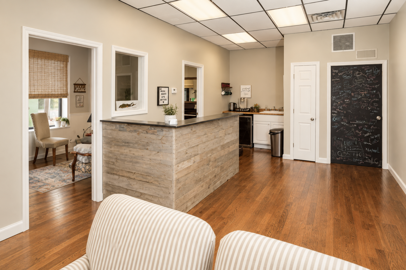 Interior view of small lounge or waiting area with wooden floor, partial view of beige and white striped armchair, adjacent to a kitchen area with a wooden counter, white cabinets, a small sink, and a chalkboard wall with colorful writing on the right side.