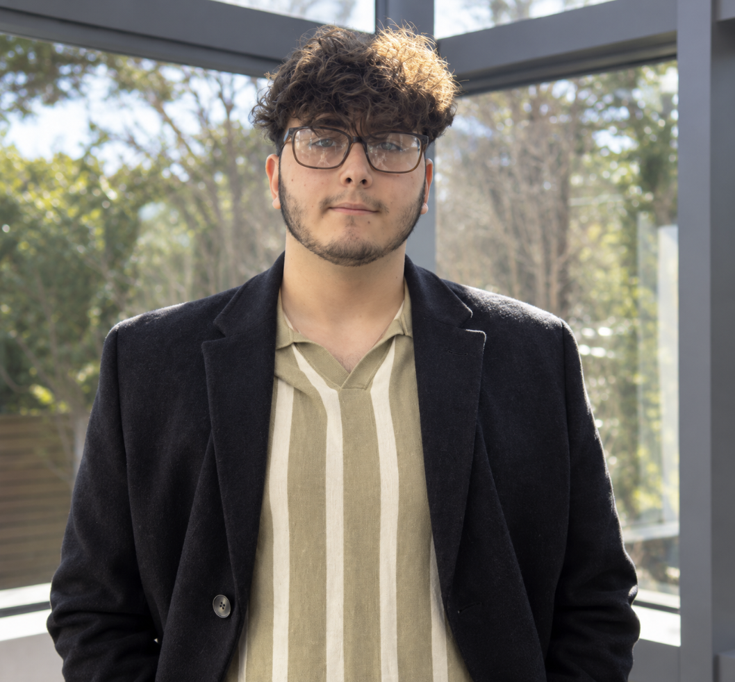 Young man with curly hair and glasses standing outdoors in front of glass windows, wearing a dark blazer over a beige shirt with vertical stripes.