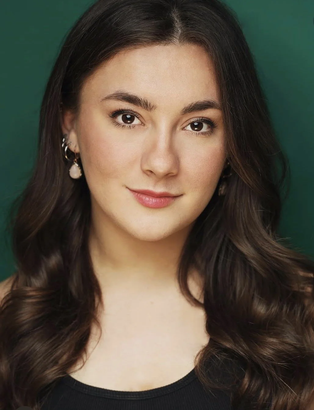A young woman with long, dark, wavy hair, light skin, and wearing earrings, looking directly at the camera with a slight smile against a green background.