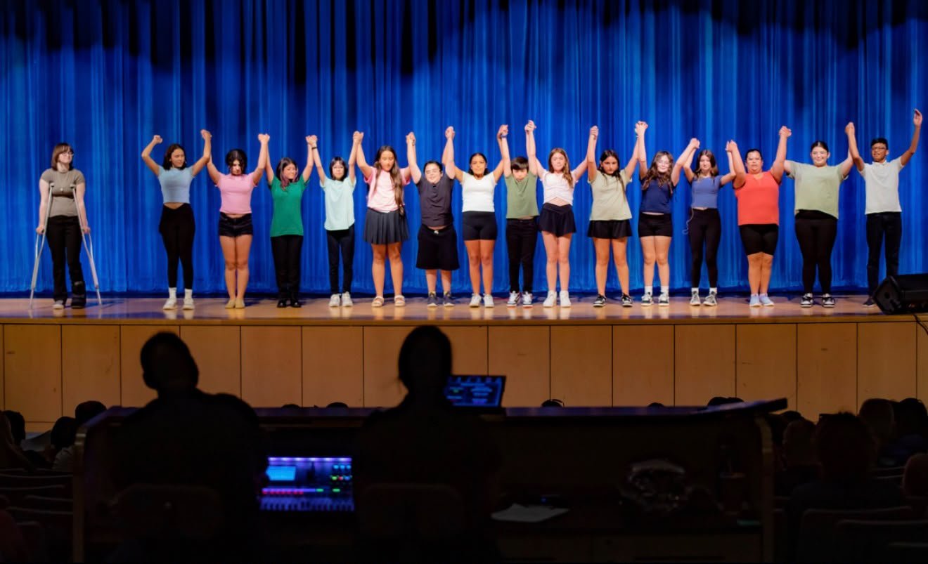 Group of children performing on stage with blue curtain backdrop, some with raised fists, and an audience watching.