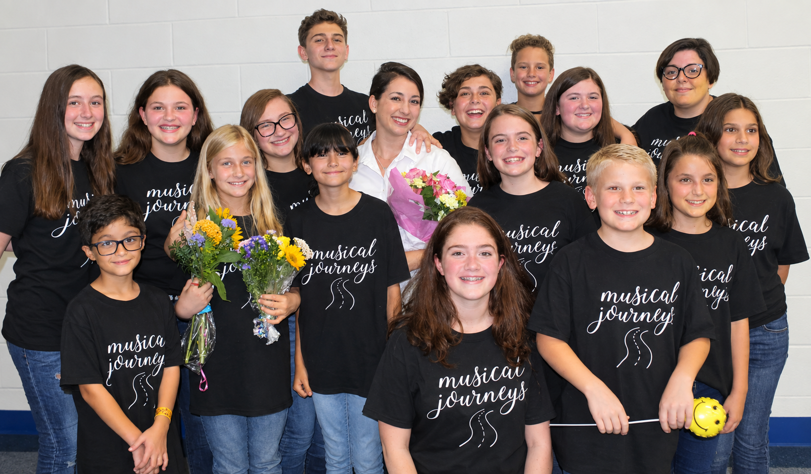 Rebecca Cypher and her 16 students standing holding flowers and wearing the t-shirts with a musical journeys logo.