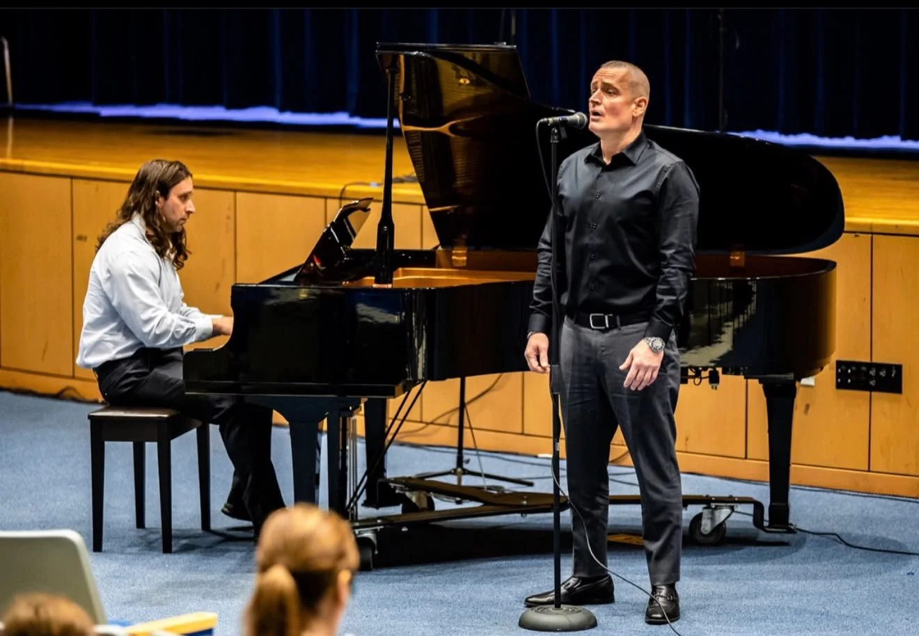 A man singing into a microphone on stage with a pianist playing a grand piano beside him during a performance.