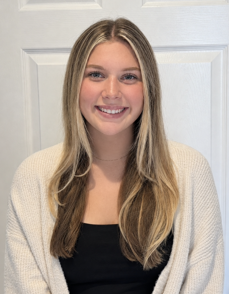 Young woman with long blonde hair, blue eyes, smiling, wearing a black top and cream-colored cardigan, standing in front of a white paneled door.