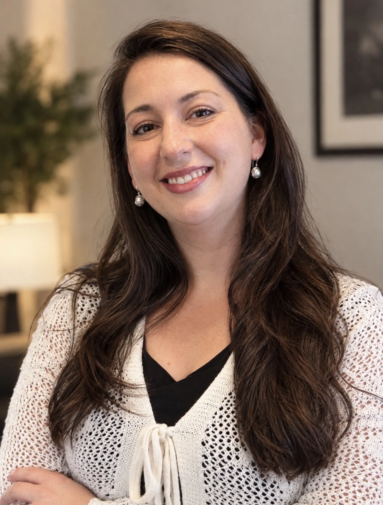 A woman with long, wavy brown hair wearing pearl earrings and a white crocheted top standing in a cozy indoor setting.