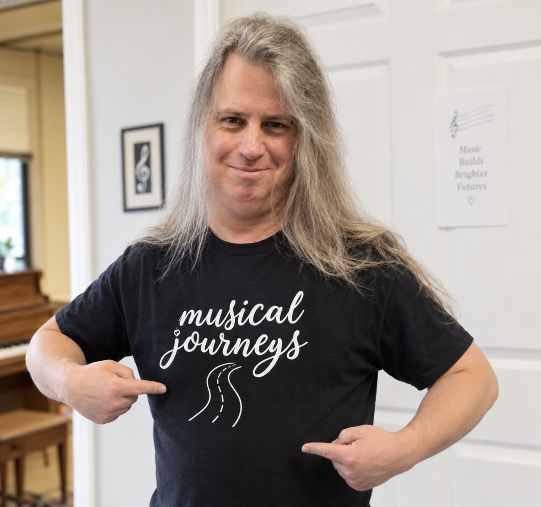 A man with long gray hair smiling and pointing to his black T-shirt that reads 'musical journeys' with a road illustration, standing indoors near a piano and framed artwork.