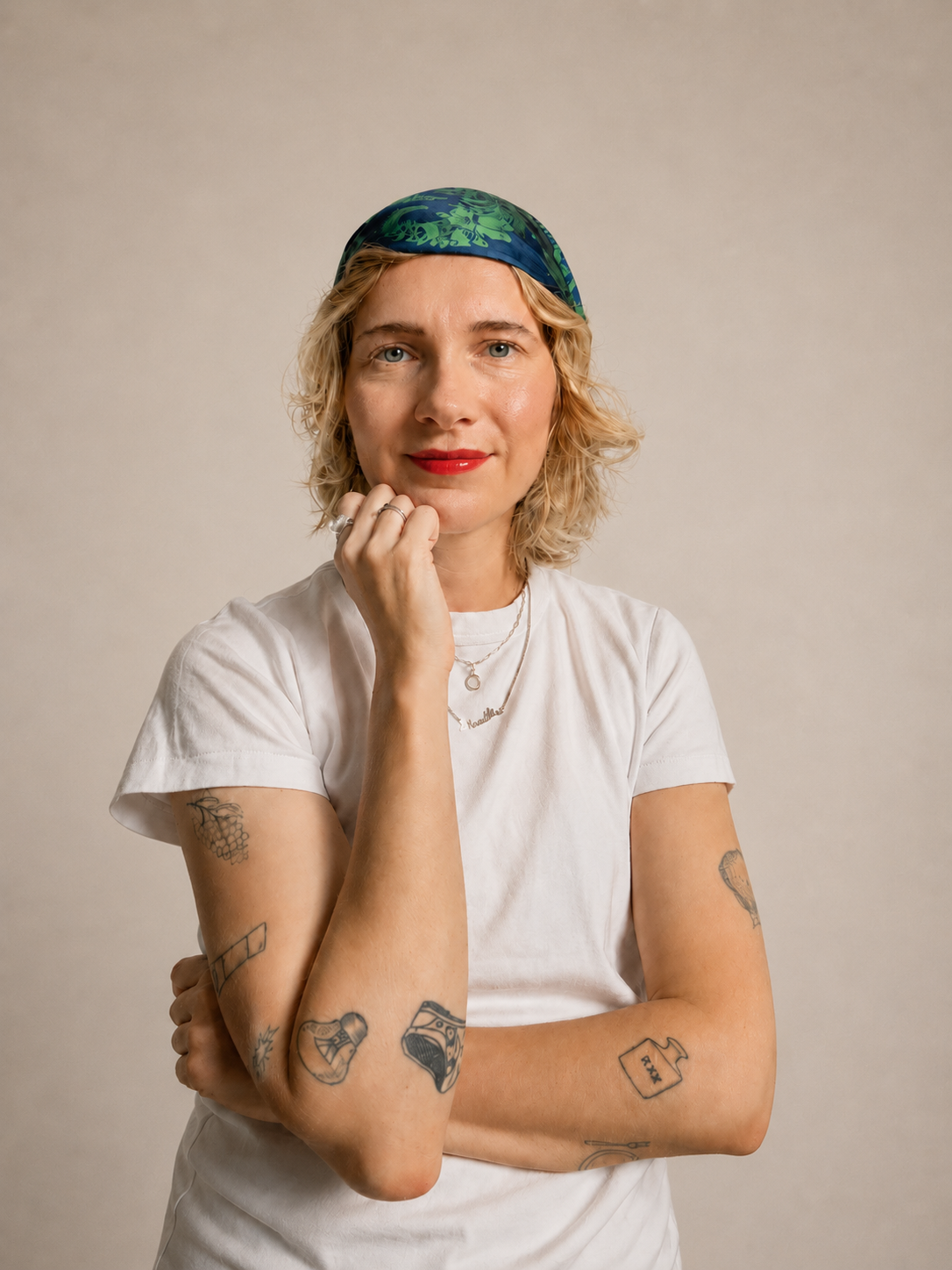 Junge Frau mit Tattoos, blondem, lockigem Haar, rotem Lippenstift, weiße T-Shirt, blauer und grüner Kopfbedeckung, posaunt nach vorne, hält Kinn mit Hand, vor neutralem Hintergrund.
