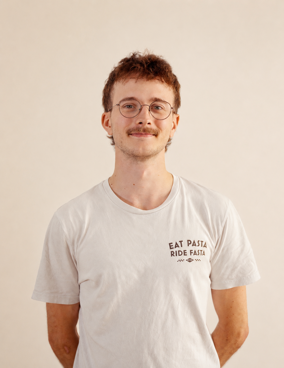 Ein junger Mann mit Brille, lockigem braunem Haar und leichten Sommersprossen trägt ein weißes T-Shirt mit dem Slogan "Eat Pasta RIDE FASTA" vor einem neutralen, hellen Hintergrund.