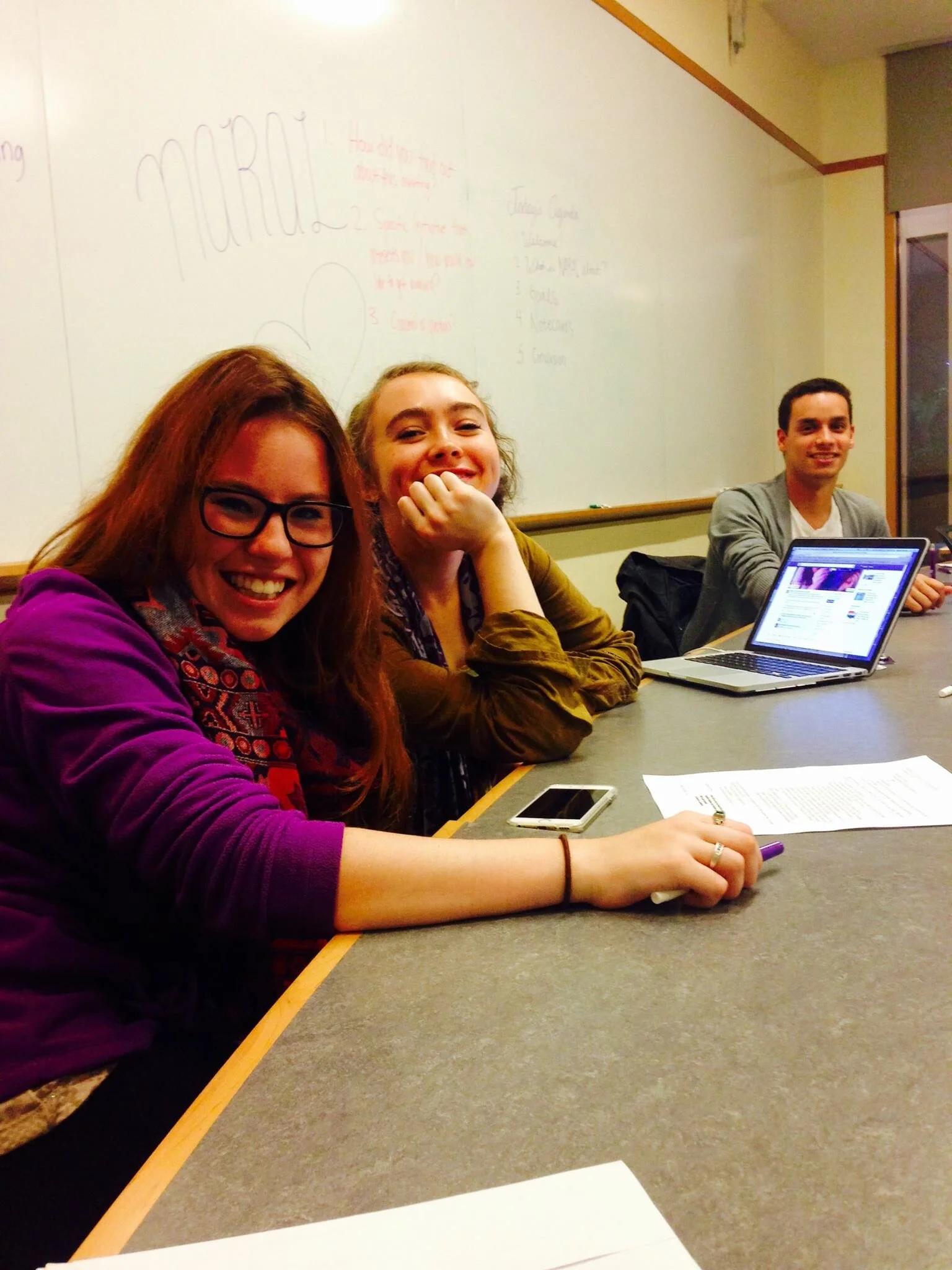 Three young adults sitting at a table in a classroom, smiling and looking at the camera, with laptops, a smartphone, and papers on the table.