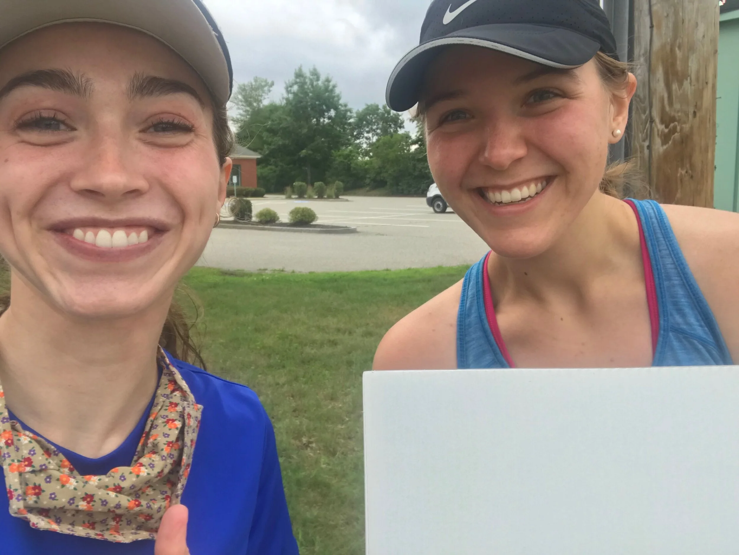 Two women smiling and taking a selfie outdoors, one wearing a beige hat and blue shirt with a floral neck gaiter, the other in a black cap and blue athletic tank top holding a blank white sign, with a parking lot and trees in the background.