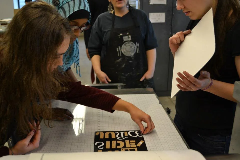 Group of five women examining a black and gold packaging label on a white grid table, with one woman pointing at it, in a workshop or studio setting.