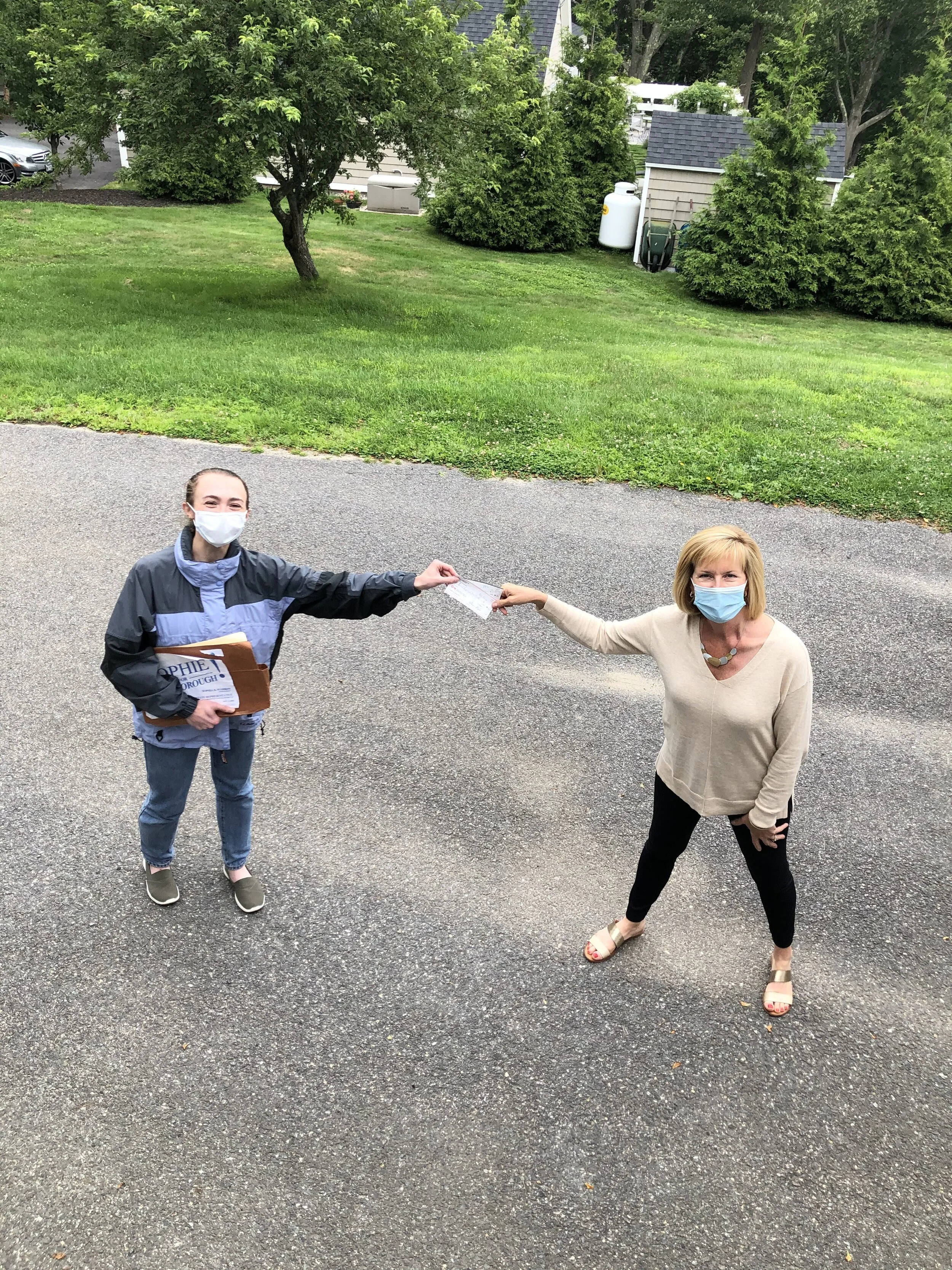 A woman and a young girl wearing masks are outdoors on a paved area, exchanging a document. The woman is dressed in a beige sweater, black pants, and sandals, while the girl is wearing a blue jacket, jeans, and sneakers. The girl is holding folders a