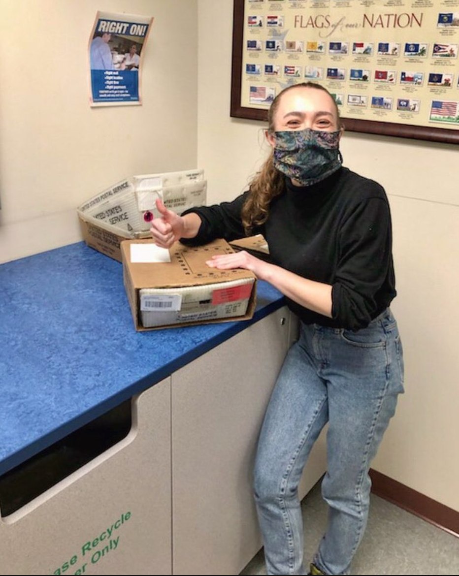 A woman wearing a face mask and casual clothes is giving a thumbs-up while standing at a table with a cardboard box. There is a poster on the wall with flags and a sign that says 'Right On!'. A reusable bin labeled 'Use Recycle Only' is beneath the t