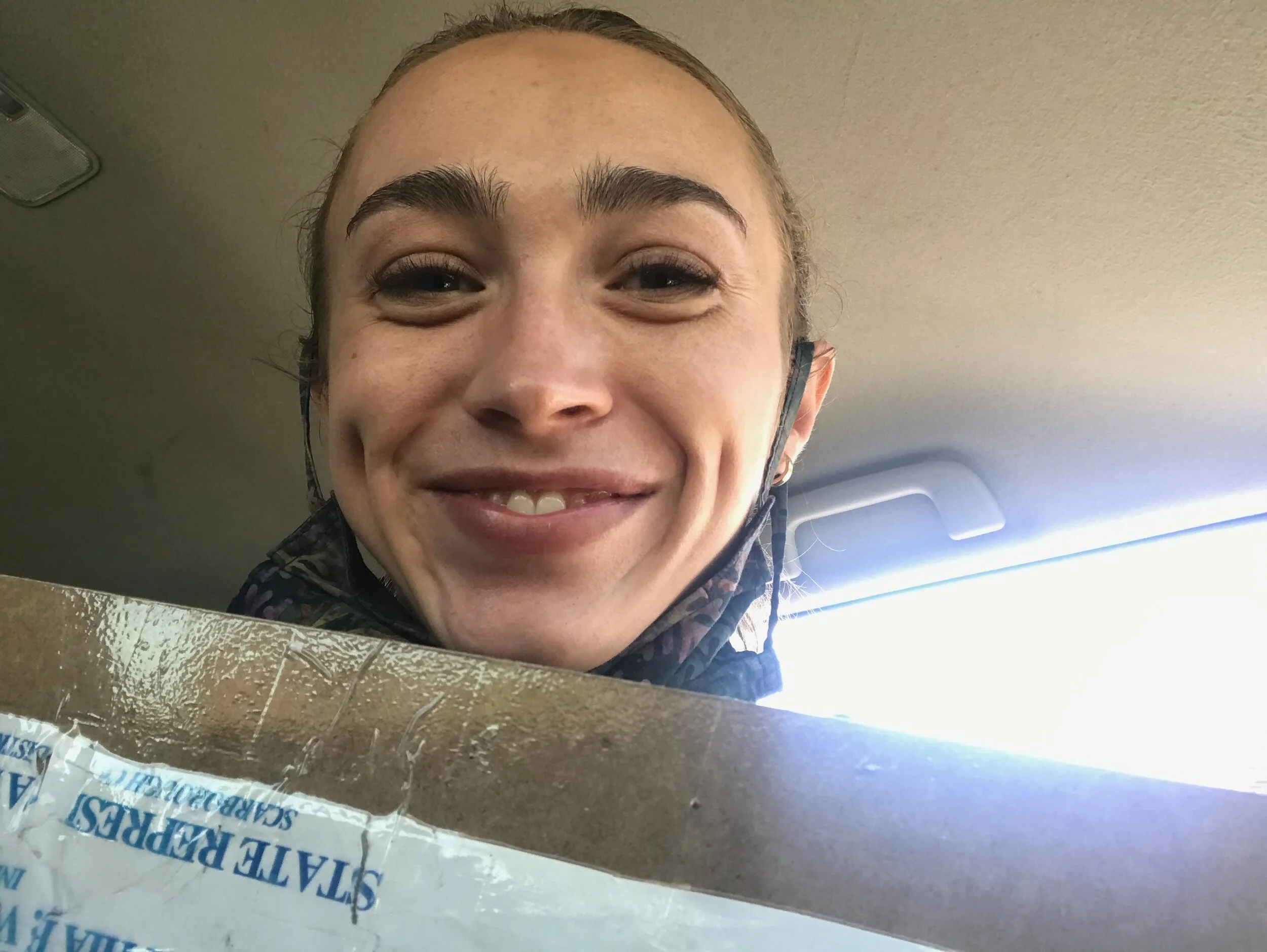 A woman smiling inside a car, wearing a face mask pulled down below her chin, with sunlight coming through the window.