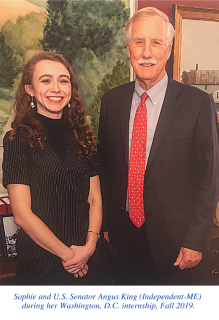 A young woman, Sophie Warren, and U.S. Senator Angus King standing together indoors, smiling. The background features a landscape painting of trees and a house during her Washington, D.C. internship, Fall 2019.