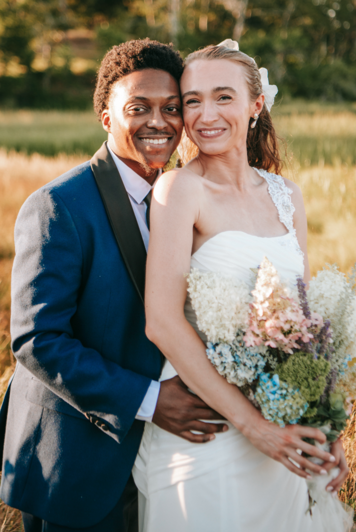 A happy couple dressed in wedding attire, standing outdoors in a field with trees in the background. The bride is holding a colorful bouquet and smiling, while the groom is embracing her from the side, both smiling at the camera.