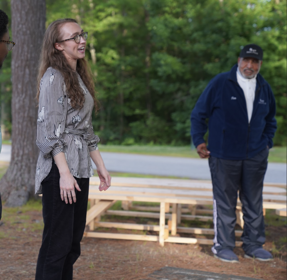 A group of people standing outdoors in a wooded area, with green trees in the background. One woman with glasses and long hair is smiling, and a man in a blue jacket and cap is also present.