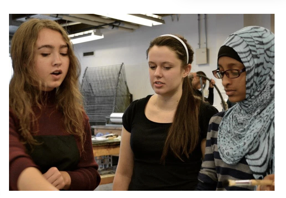 Three young women in a workshop or classroom, listening attentively to a discussion or demonstration.