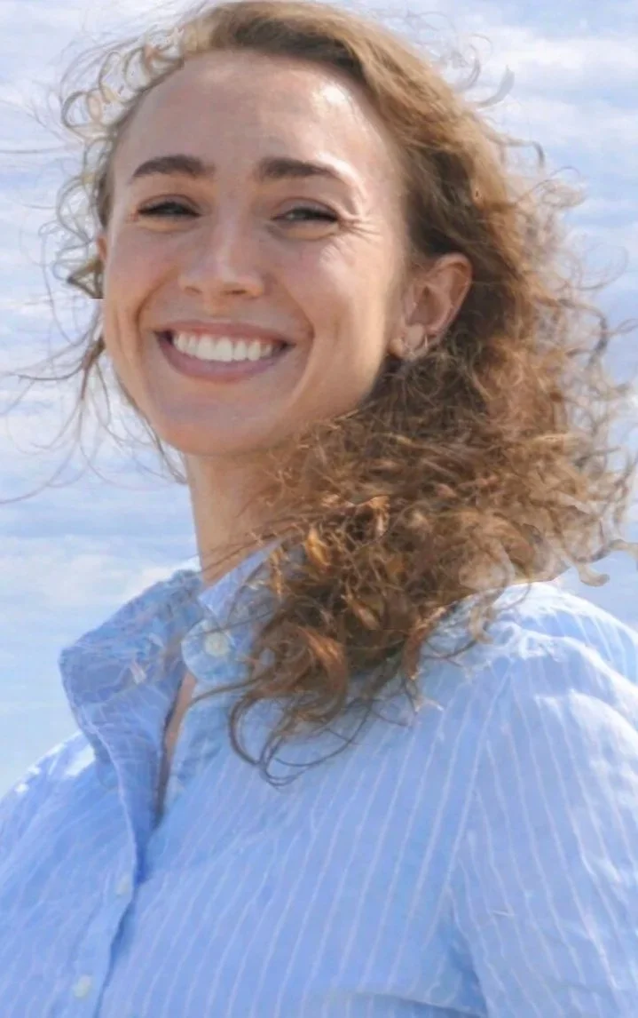 A woman named Representative Sophie Warren with curly brown hair and a big smile, wearing a light blue button-up shirt, outdoors with a cloudy sky in the background.
