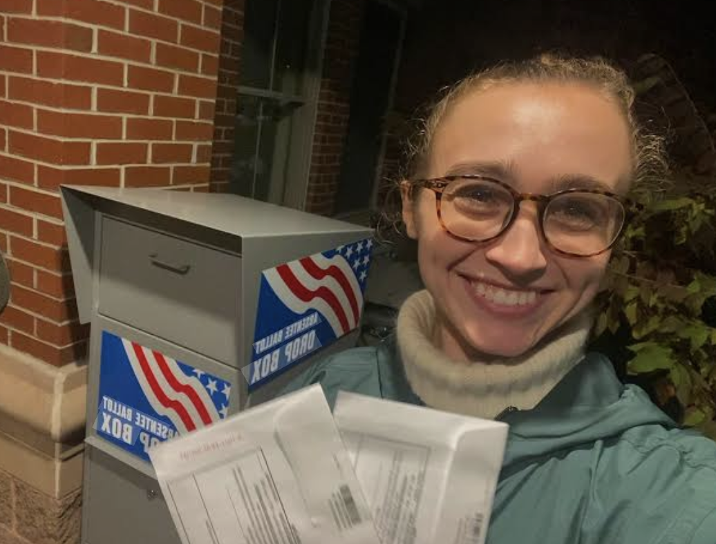 A smiling woman with glasses taking a selfie next to a mailbox with American flag decals and a package in her hand, outside a brick building at night.