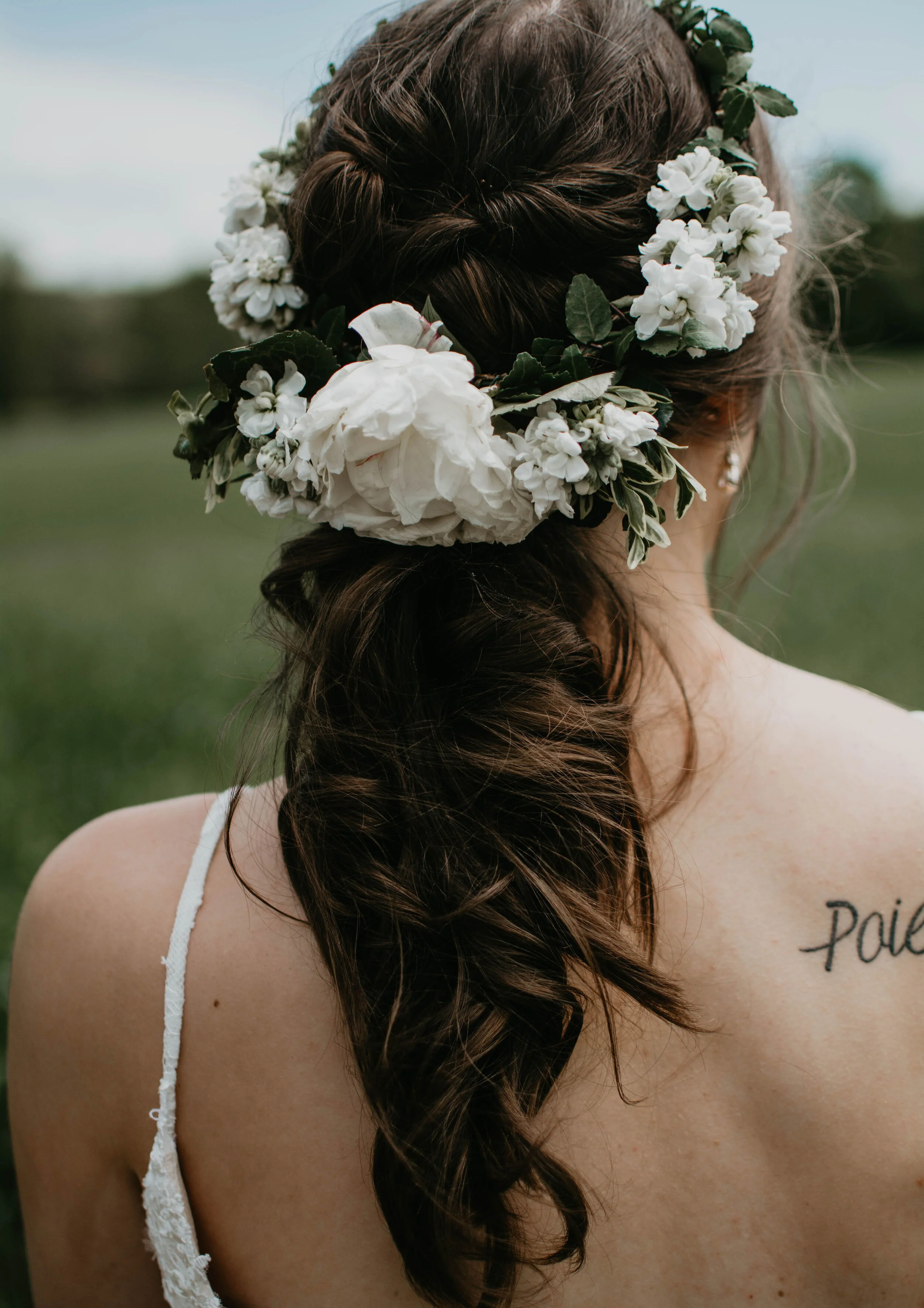 Bride with long brown hair, adorned with a white floral hairpiece, wearing a white dress with a visible tattoo on her shoulder.