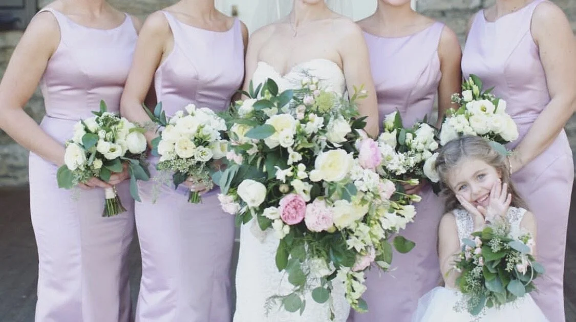Bridesmaids in lavender dresses and a bride holding bouquets, with a smiling flower girl.