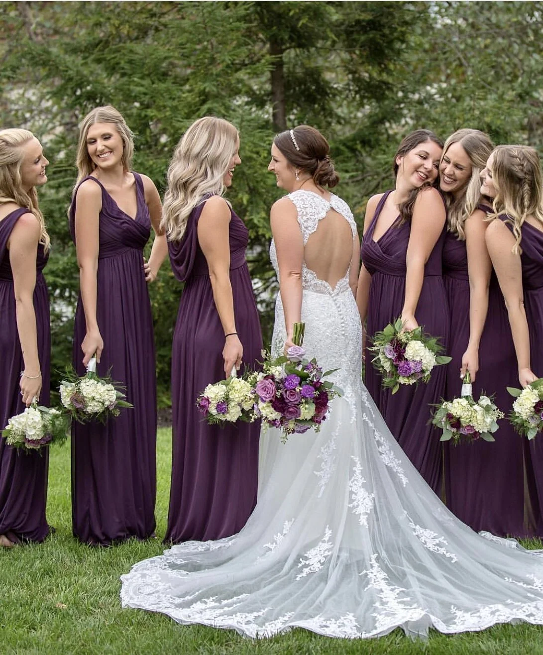 Bridal party with bride in white lace gown and bridesmaids in purple dresses holding bouquets, standing outdoors on grass.