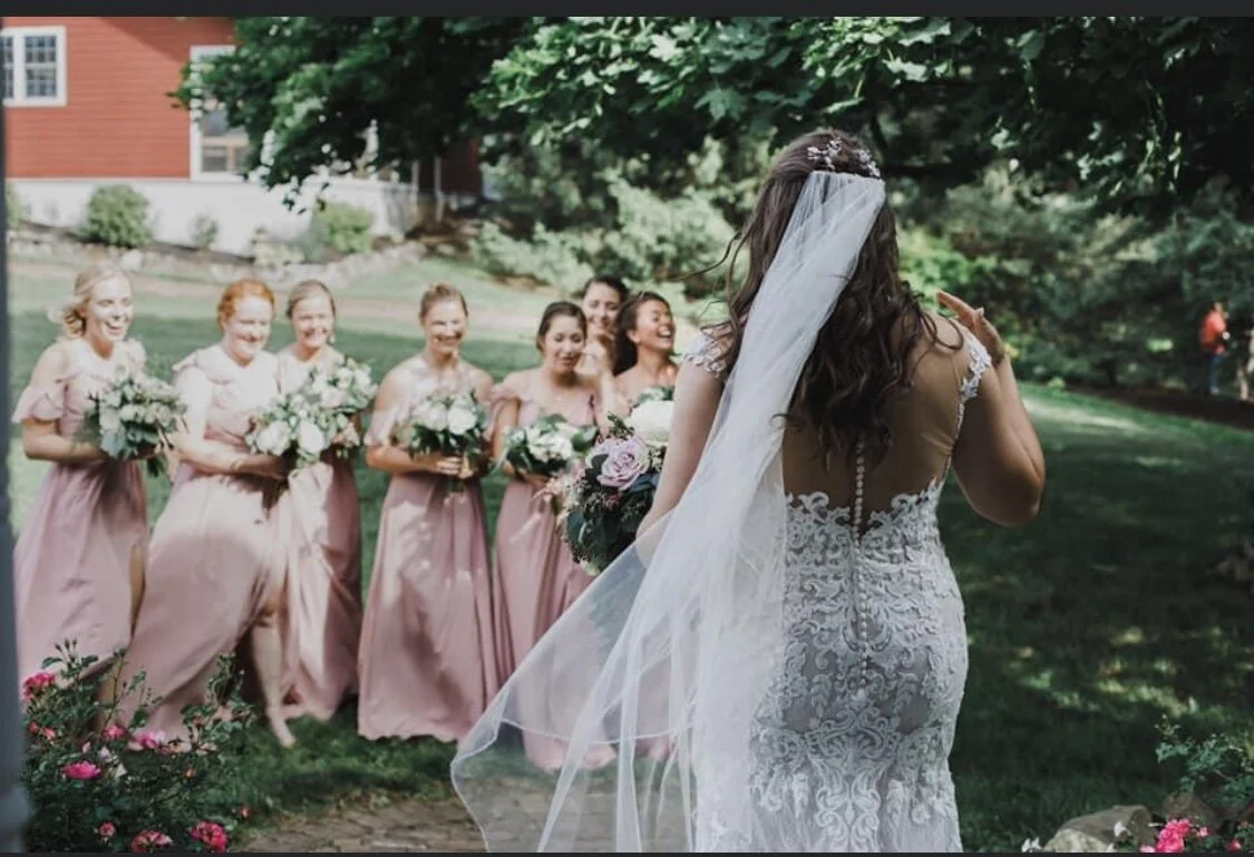 Bride in white lace dress facing bridesmaids in pink gowns outdoors.
