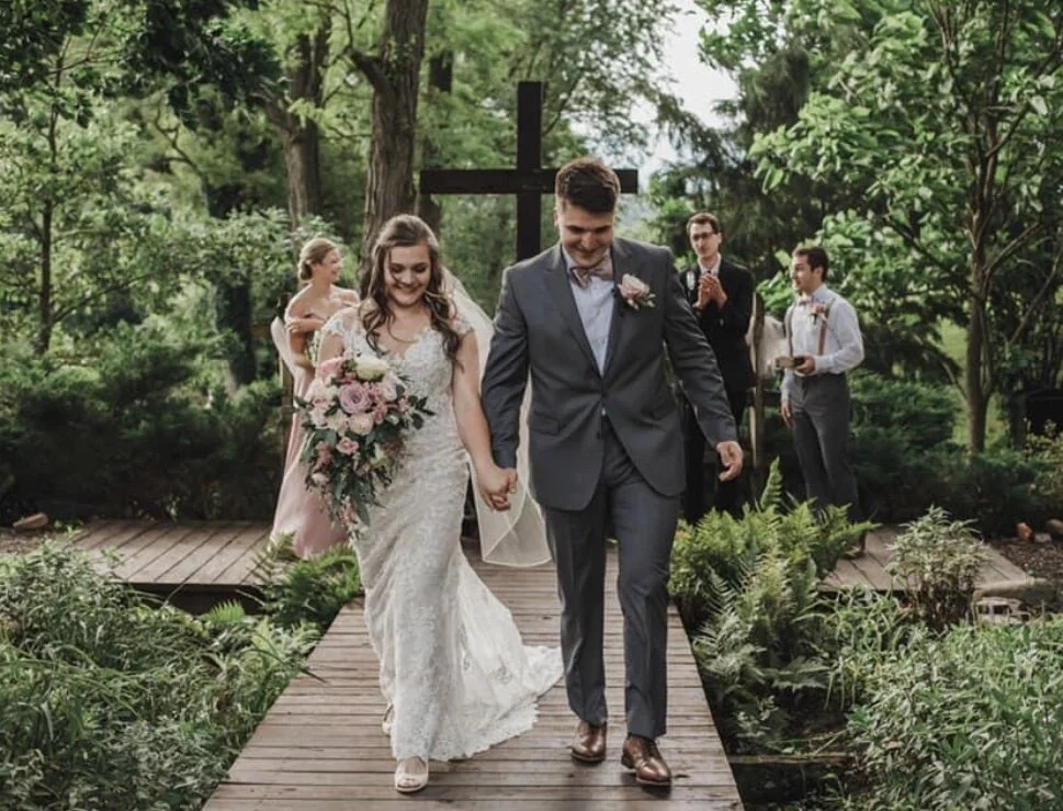 Bride and groom walking on a wooden path outdoors, surrounded by greenery, with guests in the background at a wedding ceremony.