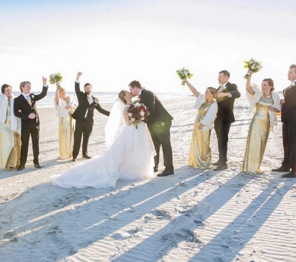 A bride and groom kissing on a sandy beach surrounded by a bridal party in gold dresses and black suits, holding flower bouquets.