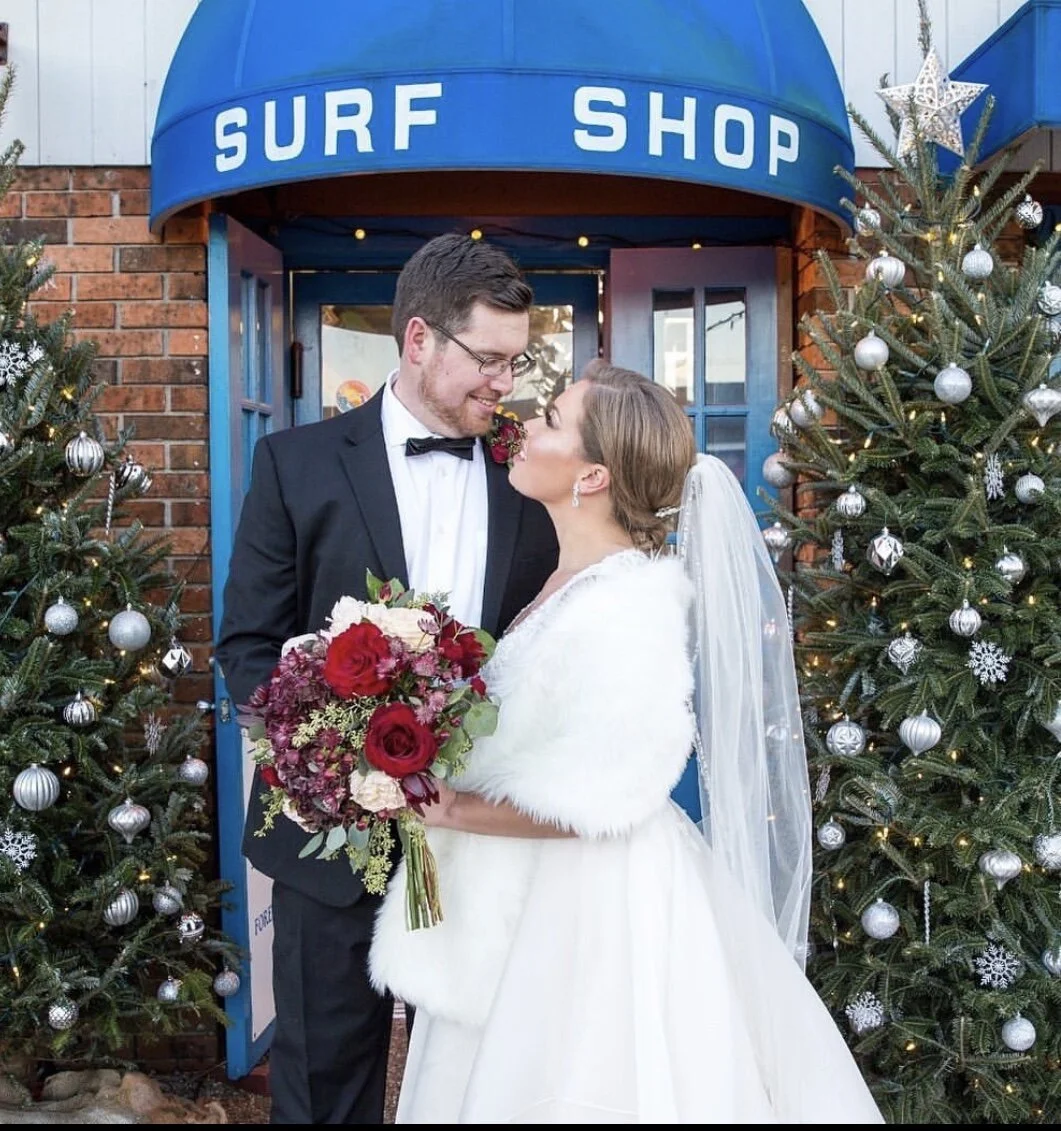 Bride in white dress and groom in black suit standing outside a surf shop, surrounded by decorated Christmas trees, holding a red and white floral bouquet.