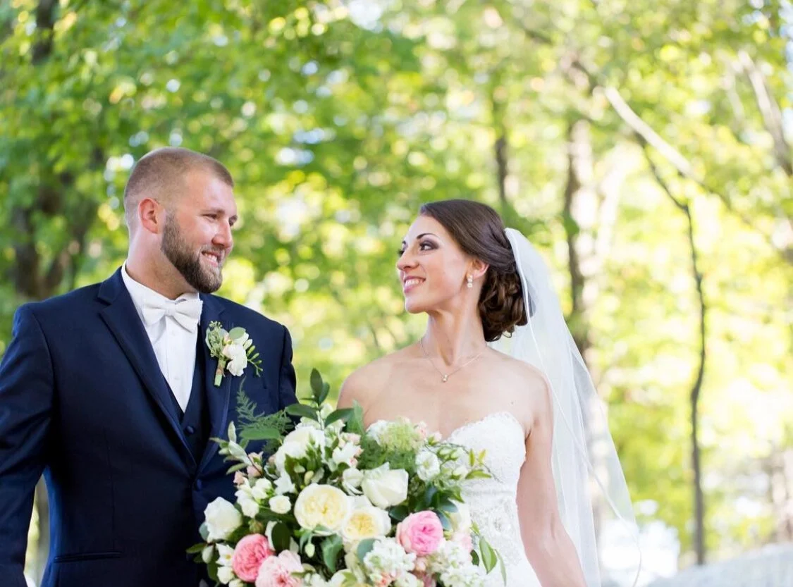 Bride and groom smiling at each other outdoors, bride holding bouquet.