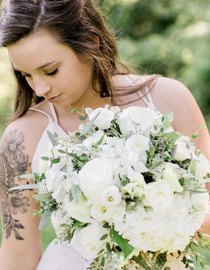Woman holding a bouquet of white flowers, with floral tattoo on her arm, in an outdoor setting.