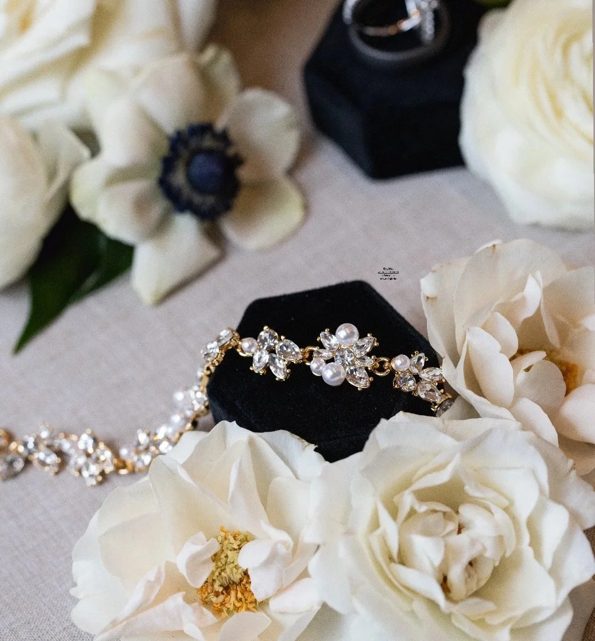 Close-up of wedding jewelry, including pearl and crystal headpiece and rings, surrounded by white flowers on a beige fabric background.