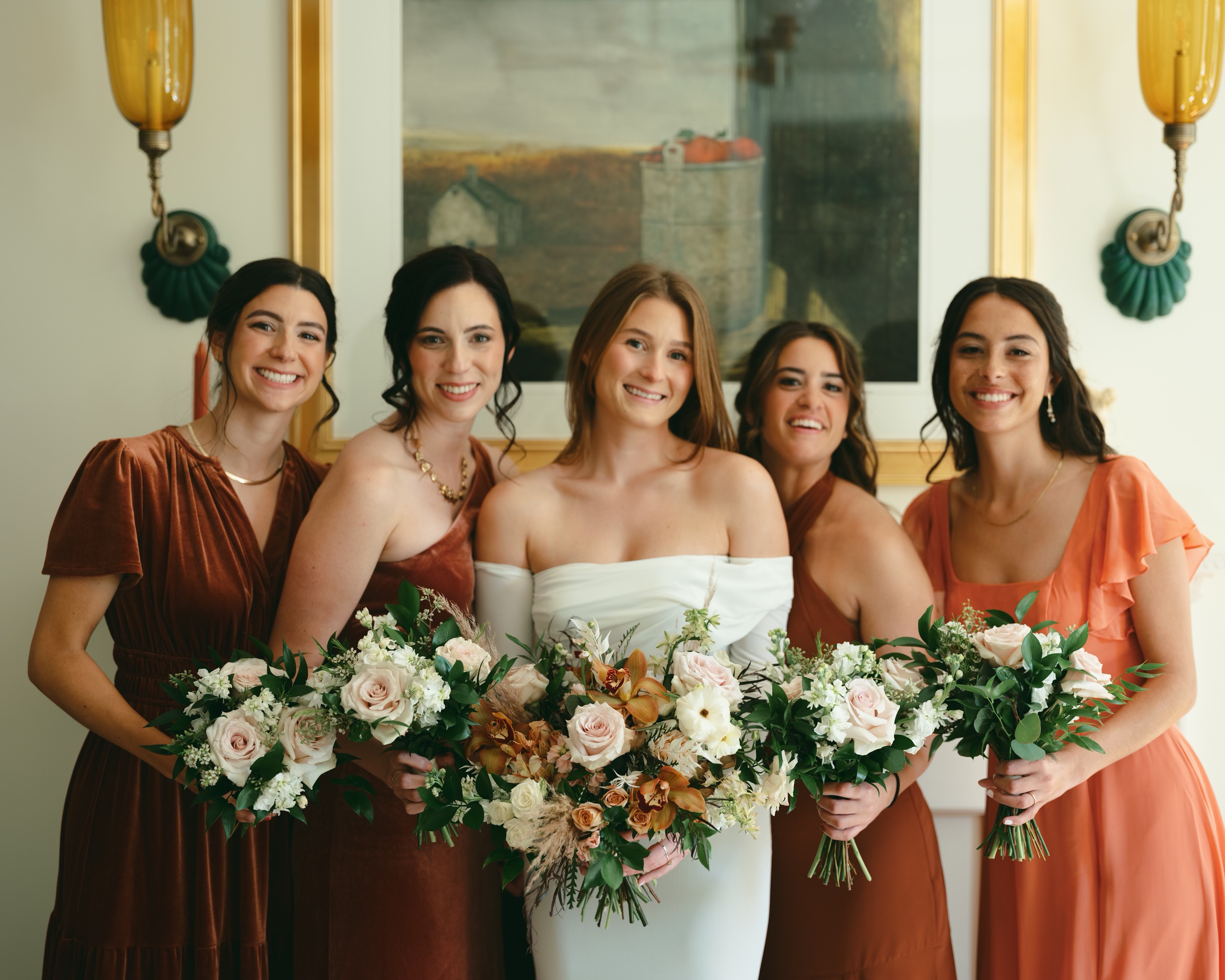 Bride with four bridesmaids holding bouquets in front of a painting, wearing elegant dresses.