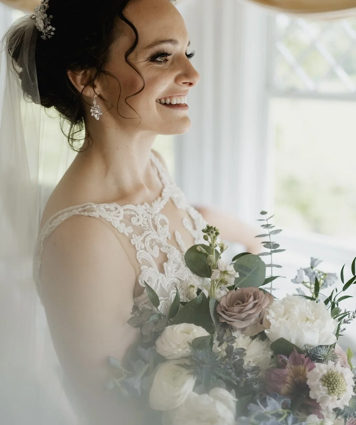 Bride smiling with bouquet, wearing lace wedding dress and veil.