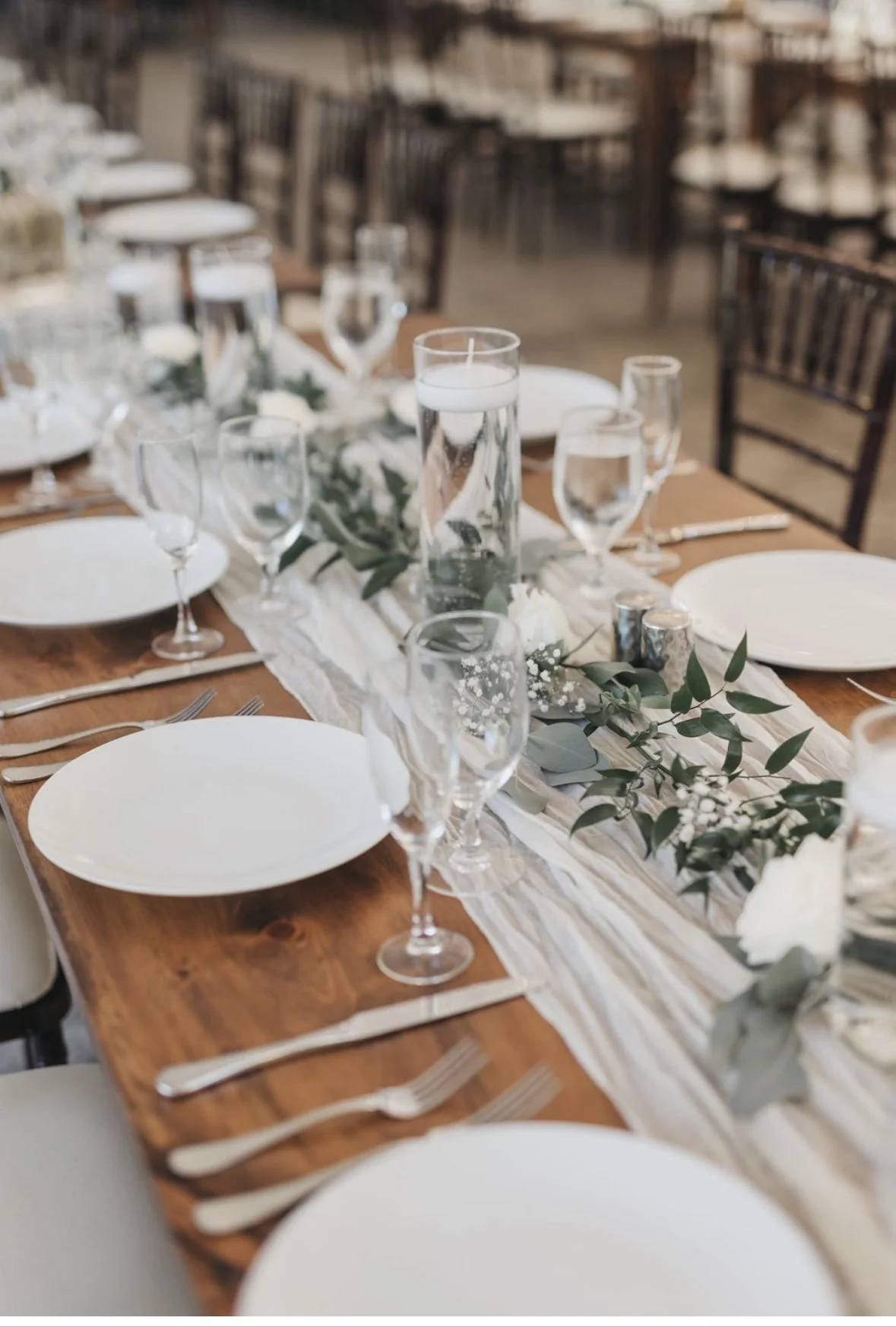 Elegant table setting with white plates, wine glasses, floating candles in vases, and greenery centerpiece on a wooden surface.