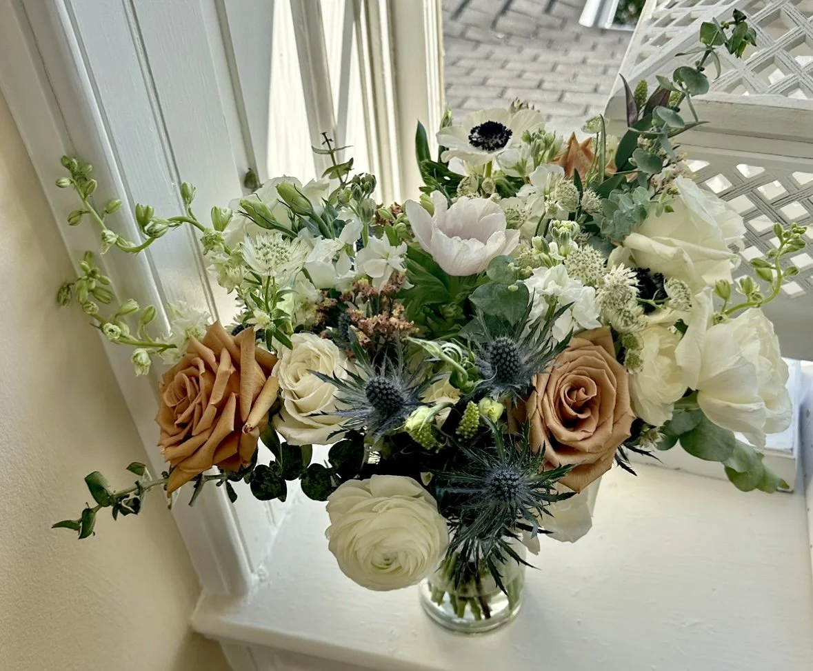 Bouquet of flowers with tan roses, white ranunculus, and greenery in a vase by a window.