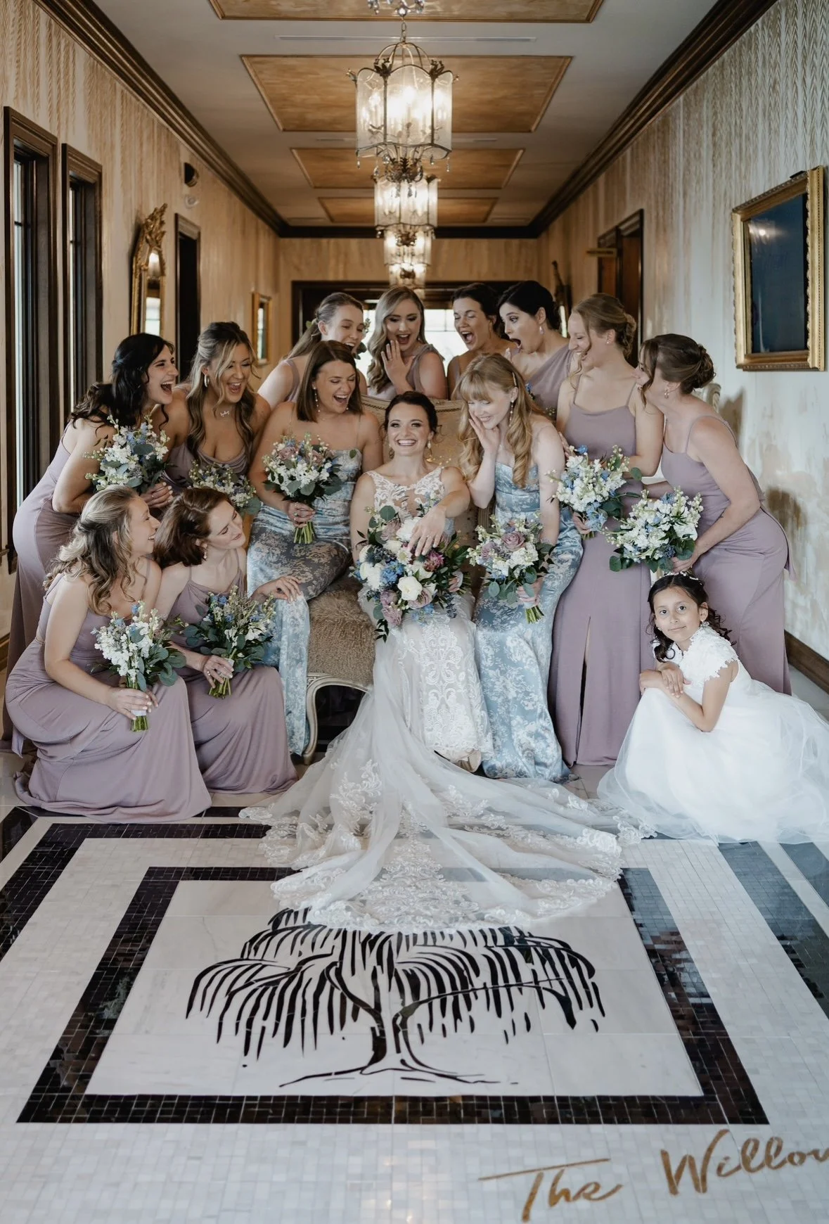 Bride with bridesmaids and a flower girl in a hallway, all holding bouquets and smiling.