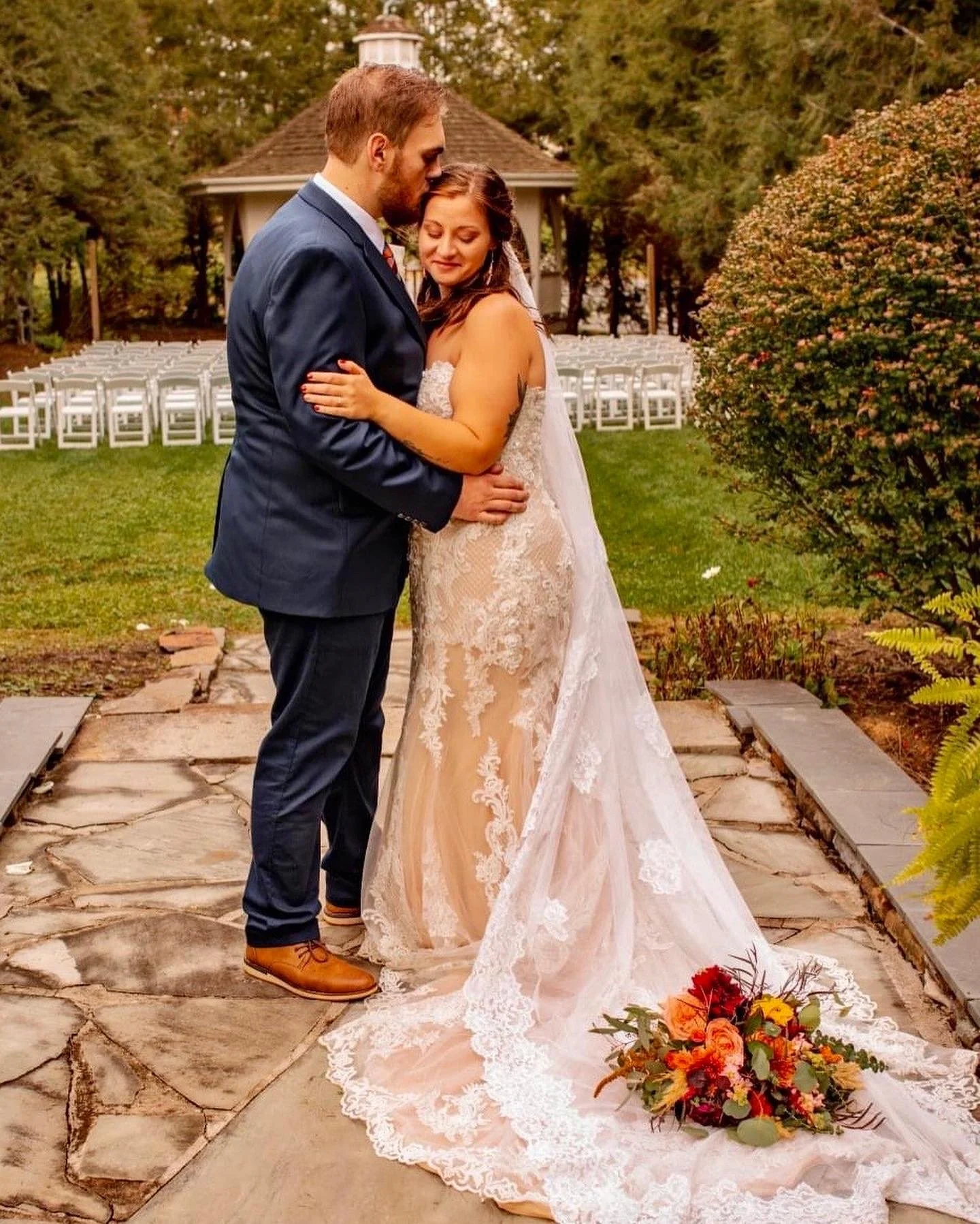 A bride and groom hugging on a stone pathway near a gazebo, with the bride wearing a lace wedding dress and veil, and the groom in a blue suit. A bouquet is on the ground next to them, and empty chairs are set up in the background.