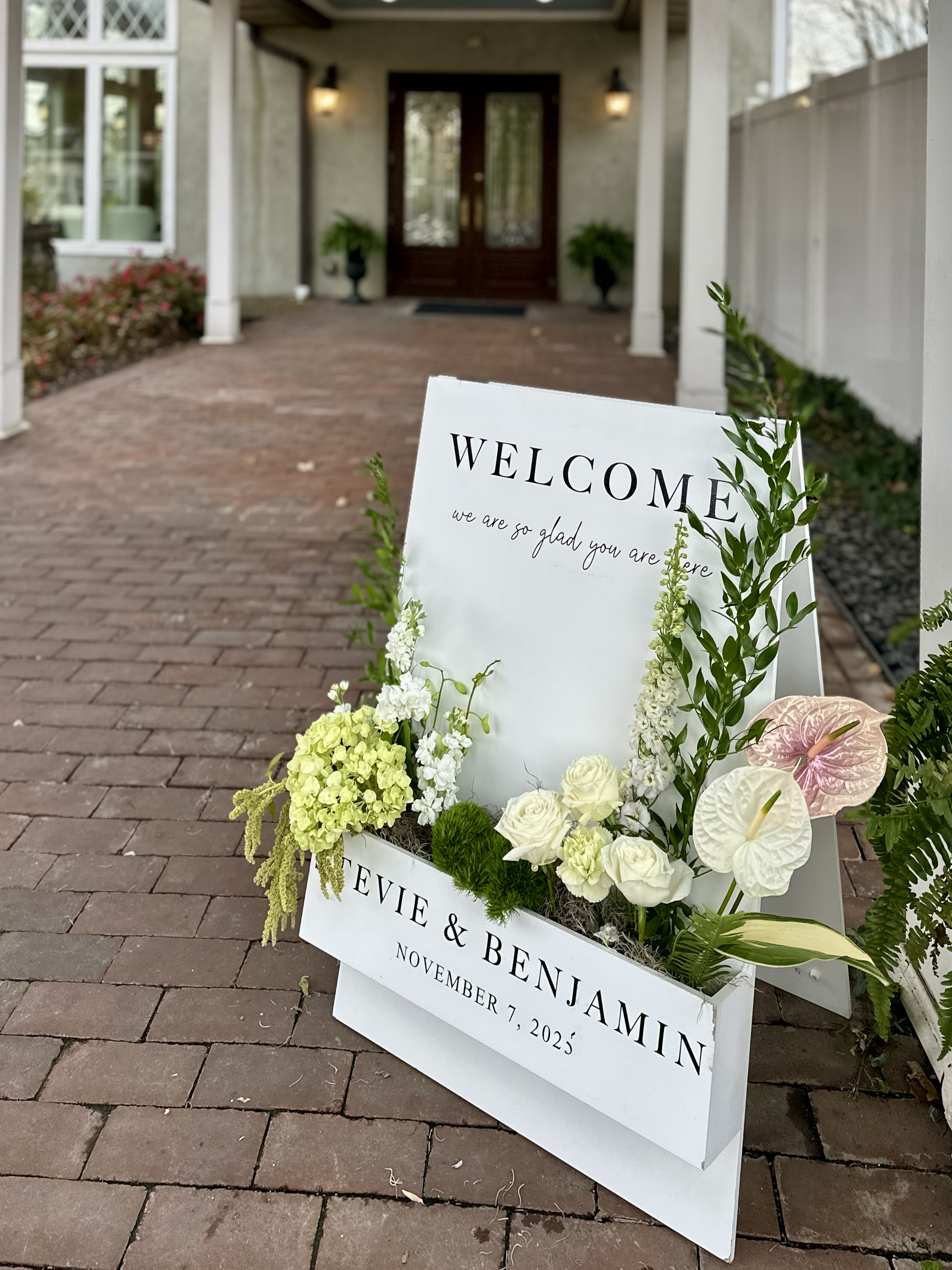 A white wedding welcome sign with anthurium, hyrgrangea, green amaranth, and greenery floral decor in a white flower box, positioned on a brick walkway outside Conservatory of Cameron Estate and Inn, PA.