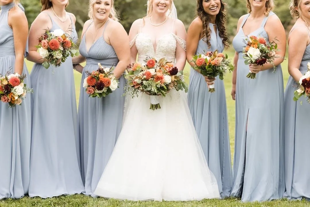 Bride with bridesmaids in matching light blue dresses holding bouquets, standing on grass.