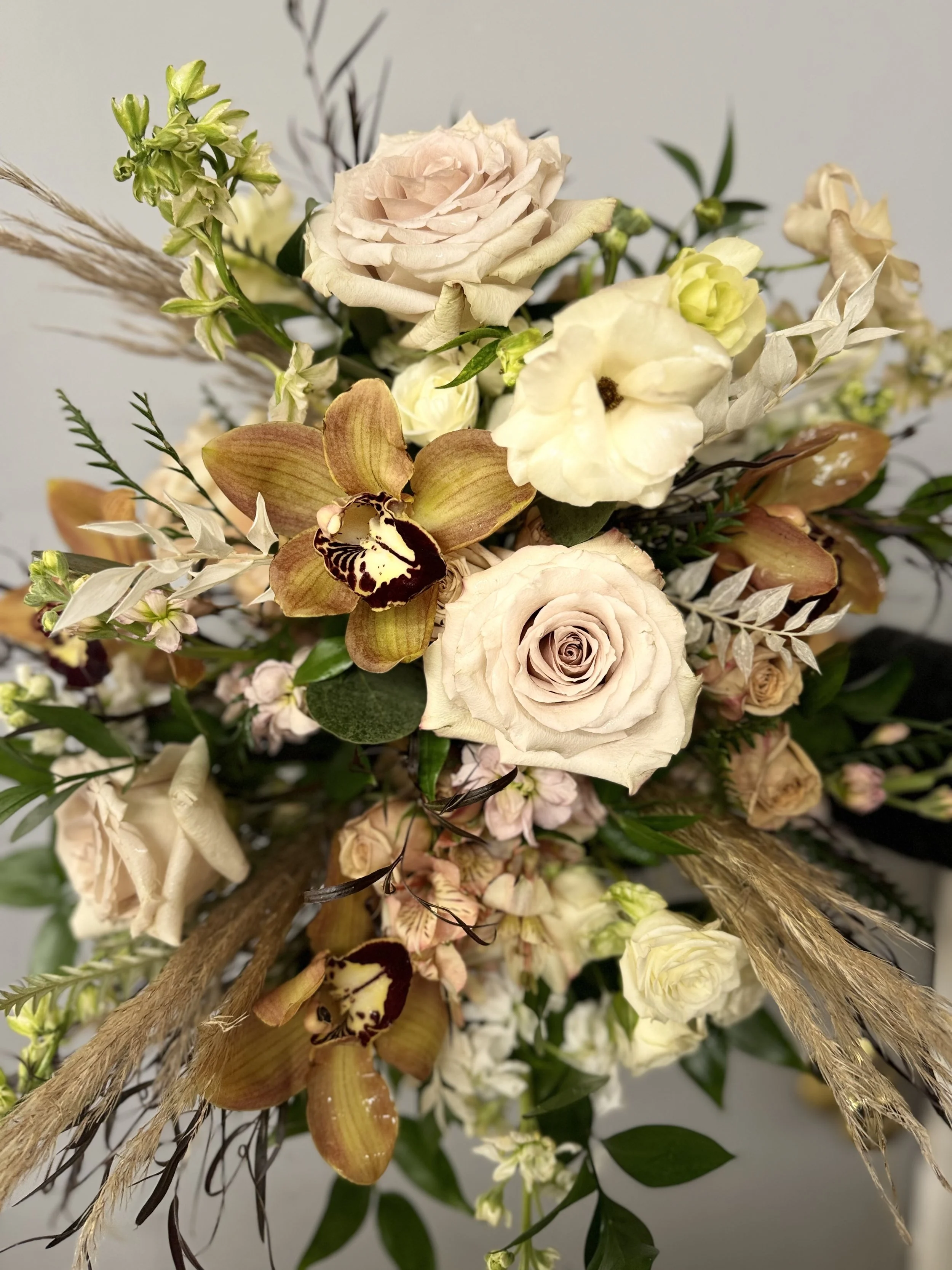 A floral arrangement featuring pale pink roses, white lisianthus, yellow and brown orchids, greenery, and decorative grasses.