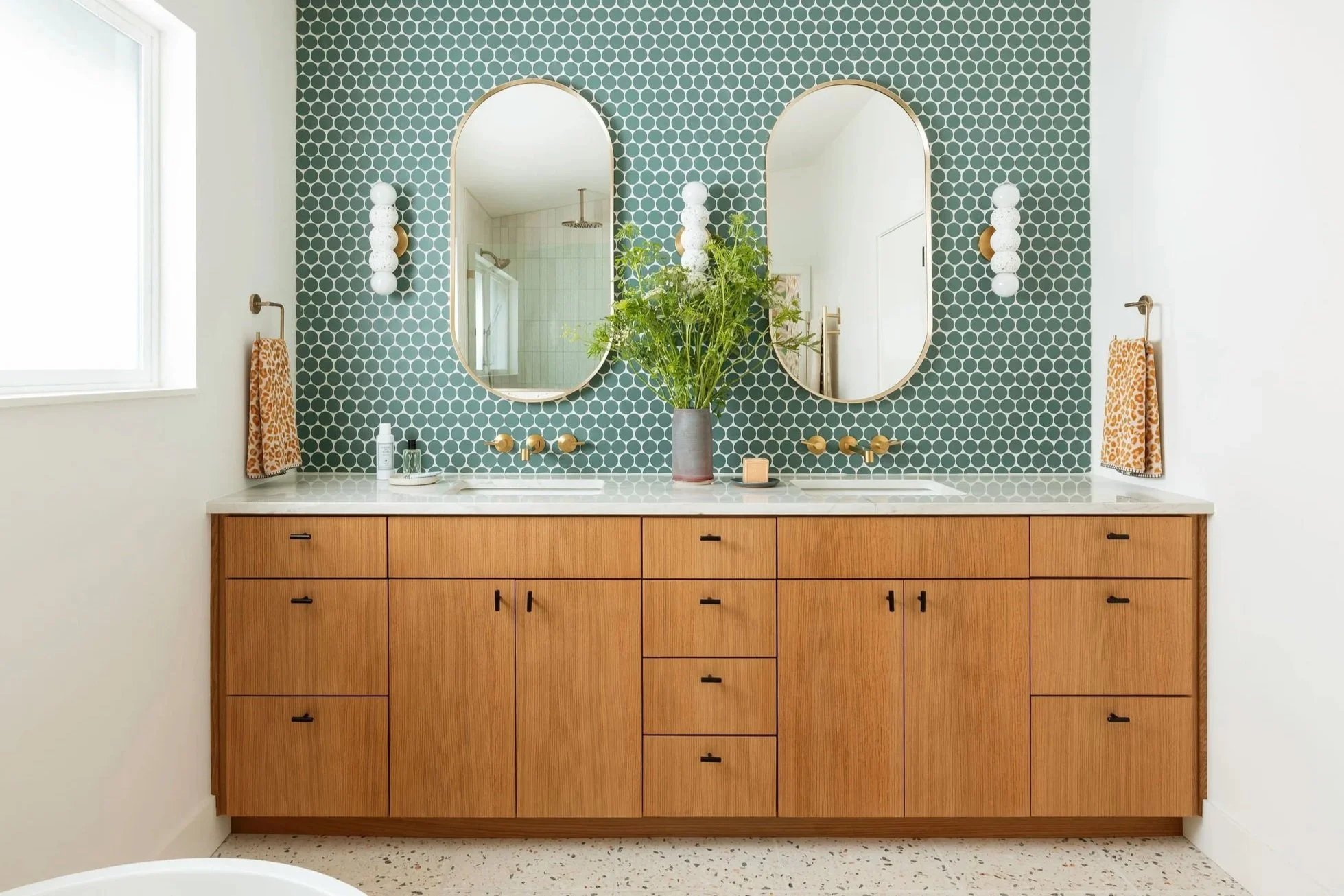 Modern bathroom with wood vanity, white countertops, double oval mirrors, green patterned tile backsplash, bouquet of flowers, and natural light.